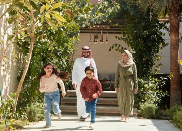 Full length front view of 6 and 7 year old siblings in casual clothing leaving shaded patio and running towards camera with smiling parents following behind.